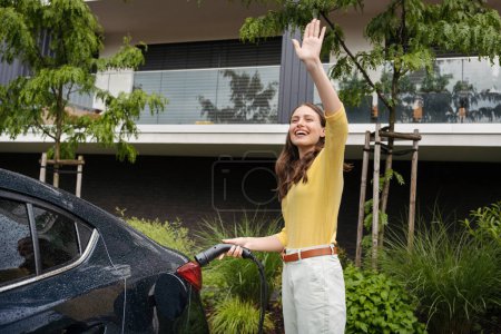 Photo for Close up of beautiful woman waving, greeting someone while charging her electric car in front of her house. Pluging in charger in charging port. - Royalty Free Image