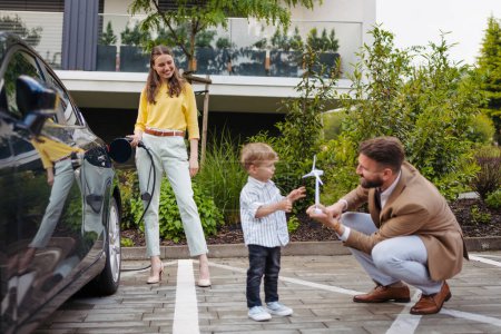 Photo for Happy family standing by beside their electric car and charging it on the street. Electric vehicle with charger in charging port. Little boy with dad playing with model of wind turbine. - Royalty Free Image
