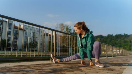 Photo for Young female runner stretching legs before her early morning run in the city. Fitness girl in sportswear preparing for evening exercise. Outdoor workout concept. - Royalty Free Image