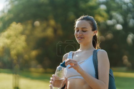 Foto de Tiro medio de gir atlética en ropa deportiva con esterilla de yoga en el hombro bebiendo de la botella de agua reutilizable. - Imagen libre de derechos