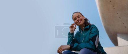 Photo for Young fitness woman listening to music with headphones, resting after workout session in the city. Beautiful sporty woman enjoying sunrise after morning excercise. Banner with copyspace. - Royalty Free Image