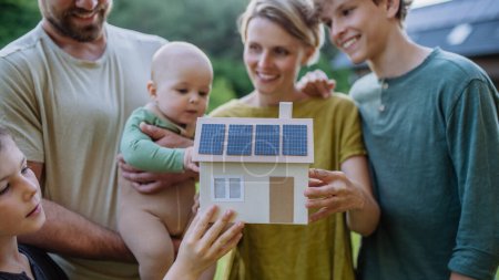 Happy family with three children holding a model of house ith solar photovoltaics. Alternative energy, saving resources and sustainable lifestyle concept.