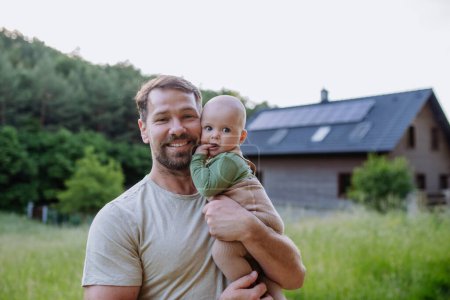 Téléchargez les photos : Père heureux avec bébé garçon debout devant leur maison avec des panneaux solaires. - en image libre de droit