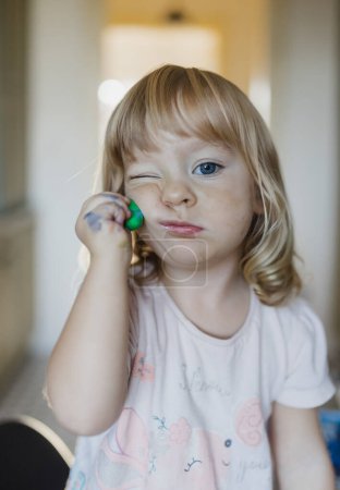 Portrait of cute little girl playing with playdough. Rolling, squashing playdough on cheek.