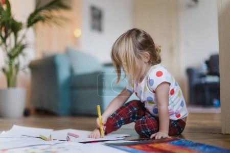 Téléchargez les photos : Mignon dessin de petite fille avec des crayons, assis sur le sol dans le salon. Décrivant son propre pied sur papier. - en image libre de droit