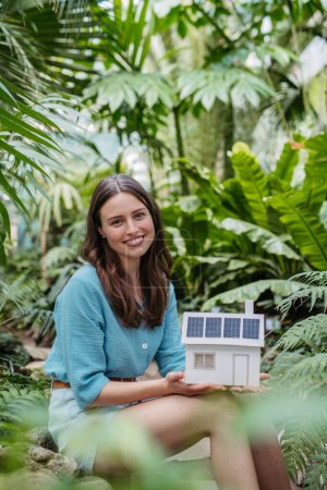 Téléchargez les photos : Jeune femme dans la jungle tenant modèle de maison avec panneaux solaires, concept d'énergie renouvelable et de protection de la nature. - en image libre de droit
