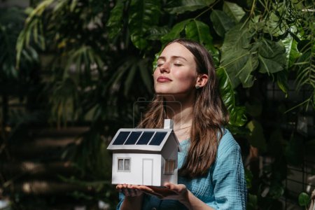 Foto de Mujer joven en selva sosteniendo modelo de casa con paneles solares, concepto de energía renovable y protección de la naturaleza. - Imagen libre de derechos