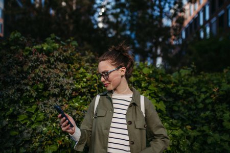 Foto de Retrato de una mujer joven desplazando su smartphone y disfrutando de la naturaleza en un parque de la ciudad. - Imagen libre de derechos