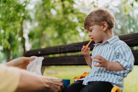 Téléchargez les photos : Mignon petit garçon mangeant des carottes hachées de la boîte à lunch, assis sur un banc du parc. Mère tenant une collation saine pour ses enfants. Manger dehors dans le parc de la ville. - en image libre de droit