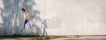 Photo for Female athlete in sportswear exercising in the city. Running in front of concrete wall casting shadow. Banner with copy space. - Royalty Free Image