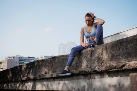 Young fitness woman in sportswear resting after hard workout session in the city. Sporty woman catching breath and drinking water after morning excercise.