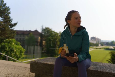 Foto de Retrato de chica atlética en ropa deportiva comiendo plátano y comprobando su teléfono inteligente después del entrenamiento. - Imagen libre de derechos
