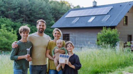 Photo for Happy family holding a model of wind turbine and paper house with photovoltaics, standing near their house with solar panels. Alternative energy, saving resources and sustainable lifestyle concept. - Royalty Free Image