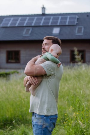 Téléchargez les photos : Père heureux avec bébé garçon debout devant leur maison avec des panneaux solaires. - en image libre de droit