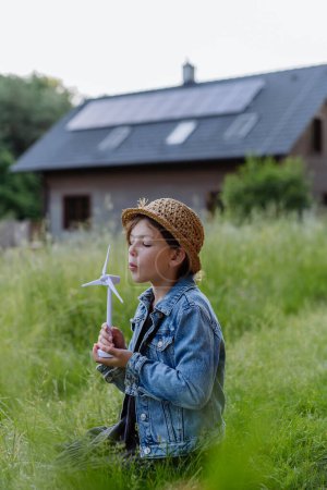 Foto de Niña sosteniendo modelo de turbina eólica, de pie frente a su casa con paneles fotovoltaicos, concepto de recursos renovables. - Imagen libre de derechos