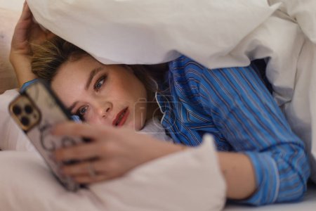 Téléchargez les photos : Jeune femme allongée sur son lit et faisant défiler un smartphone. - en image libre de droit