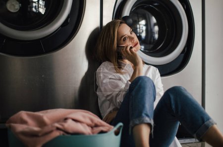 Photo for Young woman calling with smartphone, sitting in a laundry room. - Royalty Free Image
