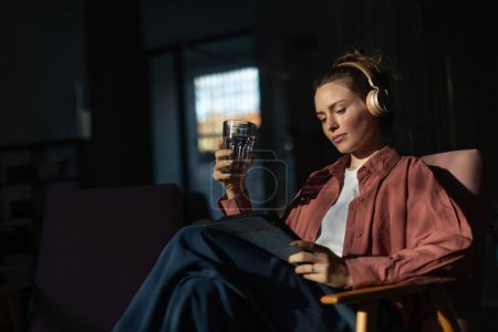 Téléchargez les photos : Jeune femme assise dans un fauteuil, écoutant de la musique et travaillant sur une tablette numérique. - en image libre de droit