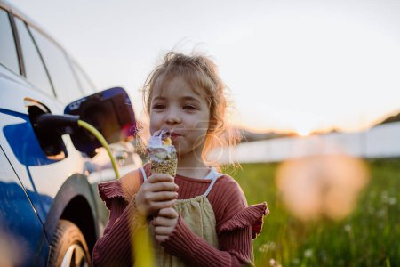 Foto de Niña comiendo un helado mientras carga su coche eléctrico. - Imagen libre de derechos