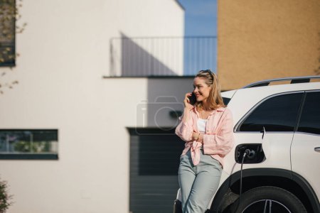Téléchargez les photos : Jeune femme appelant et rechargeant voiture électrique à la maison, concept de transport durable et économique. - en image libre de droit