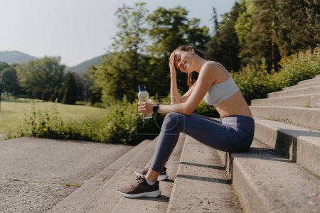 Photo for Side view of young fitness woman in sportswear resting after workout session in the city. Beautiful sporty woman drinking water on concrete stairs after morning excercise. - Royalty Free Image