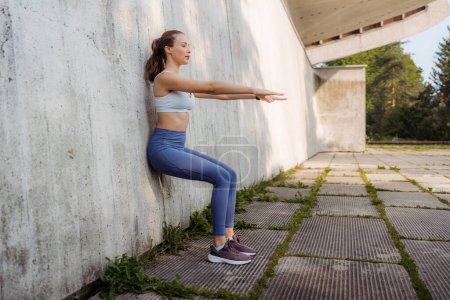 Foto de Retrato de una joven deportista en ropa deportiva haciendo ejercicio al aire libre. Mujer de fitness frente a un muro de hormigón en la ciudad. Concepto de estilo de vida saludable. Entrenamiento callejero. - Imagen libre de derechos