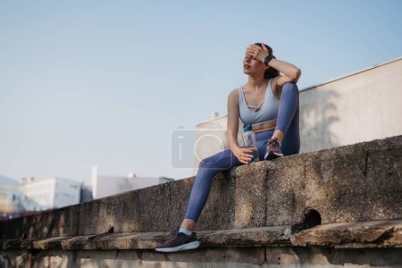 Foto de Joven mujer fitness en ropa deportiva descansando después de una sesión de entrenamiento duro en la ciudad. Mujer deportiva respirando y bebiendo agua después del ejercicio matutino. - Imagen libre de derechos