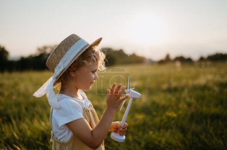 Photo for Little girl with model of wind turbine, standing in the middle of meadow Concept of renewable resources. Importance of alternative energy sources and long-term sustainability for future generations - Royalty Free Image