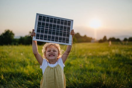 Little girl with model of solar panel, standing in the middle of meadow Concept of renewable resources. Importance of alternative energy sources and long-term sustainability for future generations