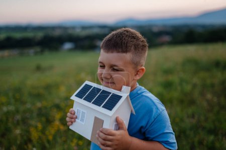 Photo for Boy with model house with installed of solar panels, standing in the middle of meadow. Concept of alternative energy, saving resources and sustainable lifestyle concept. . Importance of alternative - Royalty Free Image