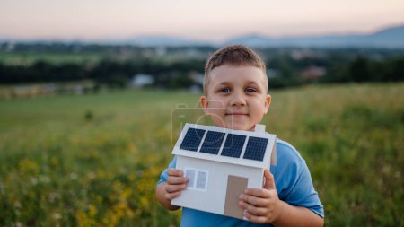Photo for Boy with model house with installed of solar panels, standing in the middle of meadow. Concept of alternative energy, saving resources and sustainable lifestyle concept. . Importance of alternative - Royalty Free Image