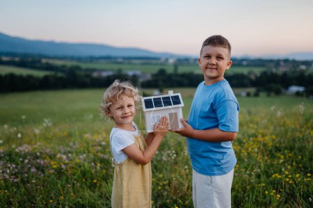 Photo for Young boy his sister holding model of house with installed solar panels. Alternative energy, saving resources and sustainable lifestyle concept. Siblings standing in the middle of meadow, representing - Royalty Free Image
