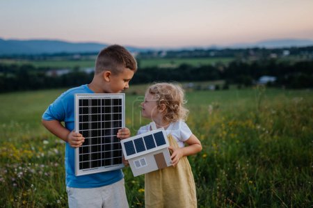 Photo for Young boy holding solar panel and his sister holding model of house with installed solar panels. Alternative energy, saving resources and sustainable lifestyle concept. Siblings standing in the middle - Royalty Free Image