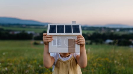 Little girl with model house with installed of solar panels, standing in the middle of meadow. Concept of alternative energy, saving resources and sustainable lifestyle concept. . Importance of