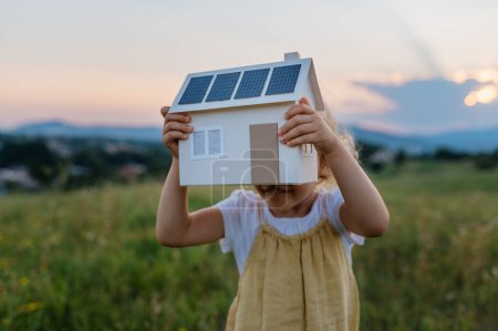 Photo for Little girl with model house with installed of solar panels, standing in the middle of meadow. Concept of alternative energy, saving resources and sustainable lifestyle concept. . Importance of - Royalty Free Image