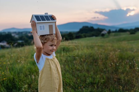 Petite fille avec maison modèle avec installation de panneaux solaires, debout au milieu de la prairie. Concept d'énergie alternative, d'économies de ressources et de mode de vie durable. . Importance de