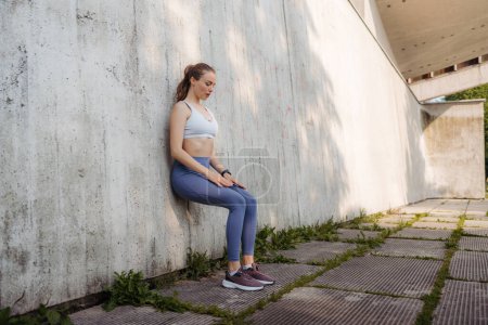Foto de Retrato de una joven deportista en ropa deportiva haciendo ejercicio al aire libre, haciendo ejercicio de sentarse en la pared. Mujer de fitness frente a un muro de hormigón en la ciudad. Entrenamiento callejero. - Imagen libre de derechos