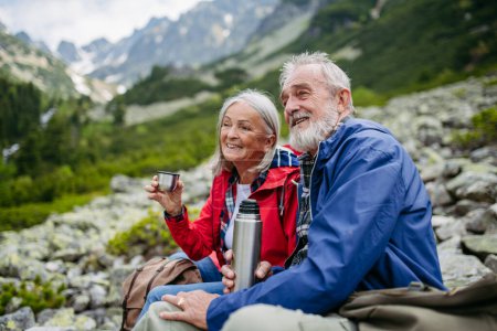 Photo for Active elderly couple hiking together in autumn mountains, on senior friendly trail. Husband and wife rehydrating, enjoying warm tea, coffee from thermos. Senior tourist with backpack getting rest - Royalty Free Image