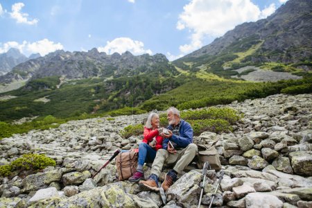 Photo for Active elderly couple hiking together in autumn mountains, on senior friendly trail. Husband and wife having healthy snack, to get energy . Senior tourist with backpacks getting rest during hike. - Royalty Free Image