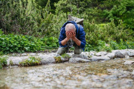 Foto de Hombre mayor activo lavándose la cara en el arroyo de montaña durante la caminata. Turista senior con mochila enfriándose por un pequeño río. - Imagen libre de derechos