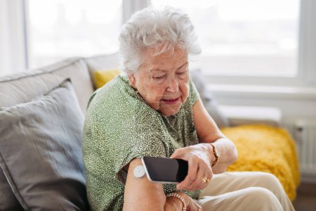 Téléchargez les photos : Patient âgé diabétique vérifiant le taux de glucose sanguin à la maison à l'aide d'un glucomètre continu. Femme âgée connectant sa MGM avec un téléphone intelligent pour voir ses niveaux de sucre dans le sang en temps réel. - en image libre de droit
