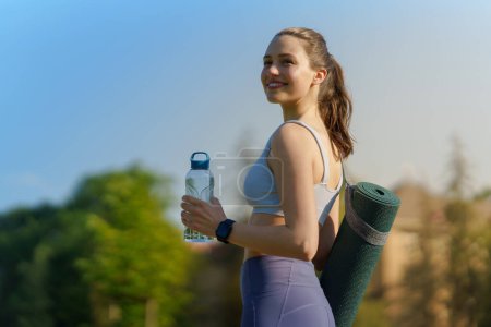 Téléchargez les photos : Vue latérale de la belle fille athlétique en vêtements de sport avec tapis de yoga sur l'épaule et la bouteille d'eau se dirigeant vers une séance d'entraînement pilates. - en image libre de droit