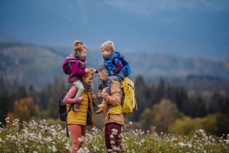 Photo for Happy parents with their little kids on piggyback at autumn walk, in the middle of colourfull nature. Concept of a healthy lifestyle. Carrying children on shoulders during the hike. - Royalty Free Image