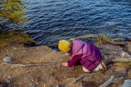 Téléchargez les photos : Petite fille jouant sur la rive du lac dans les montagnes. Touriste fille au repos près du lac, s'amuser et jouer avec des cailloux. - en image libre de droit