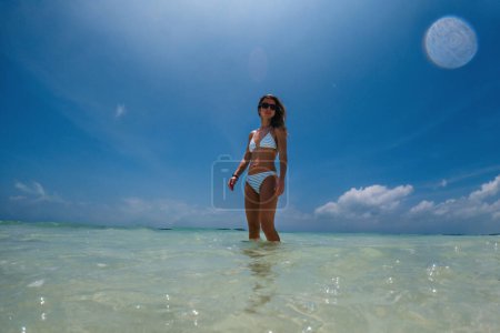 Photo for Portrait of a beautiful slim woman standing in sea water on beach in bikini. Low angle, full body shot of woman in blue swimsuit, sunglasses enjoying sandy beach and crystalline sea of Mnemba beach in - Royalty Free Image