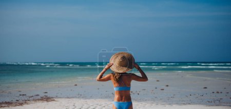 Foto de Vista trasera de una hermosa mujer delgada de pie en la playa en bikini y sombrero de paja. Cintura arriba tiro de mujer en traje de baño azul disfrutando de la playa de arena y el mar cristalino de la playa de Mnemba en Zanzíbar - Imagen libre de derechos