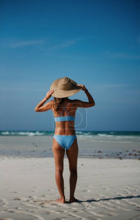 Foto de Vista trasera de una hermosa mujer delgada de pie en la playa en bikini y sombrero de paja. Foto de cuerpo completo de mujer en traje de baño azul disfrutando de la playa de arena y el mar cristalino de la playa de Mnemba en Zanzíbar - Imagen libre de derechos