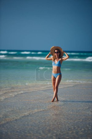 Photo for Portrait of a beautiful slim woman standing on the beach in a bikini and a straw hat. Full body shot of woman in blue swimsuit, sunglasses enjoying sandy beach and crystalline sea of Mnemba beach in - Royalty Free Image