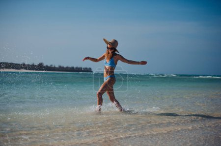 Photo for Portrait of a beautiful slim woman splashing water on the beach in a bikini and a straw hat. Full body shot of woman in blue swimsuit, sunglasses enjoying sandy beach and crystalline sea of Mnemba - Royalty Free Image