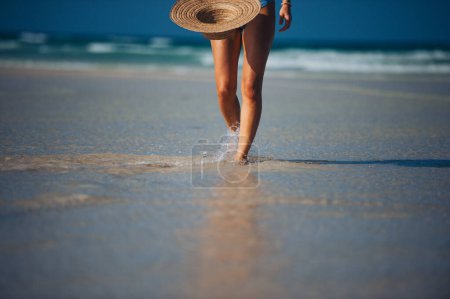 Photo for Close up of beautifuly shaped womens legs on the beach near the sea. Detail shot of woman on beach with straw hat in hand with copy space. Sandy beach and crystalline sea of Mnemba beach in Zanzibar - Royalty Free Image